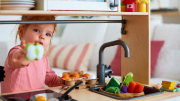 cute toddler baby girl playing on toy kitchen at home, roasting eggs and treat you with apple slice, let's share