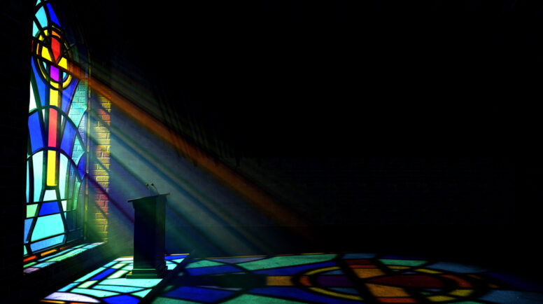 A dim old church interior lit by suns rays penetrating through a colorful stained glass window in the pattern of a crucifix reflecting colours on the floor and a speech pulpit