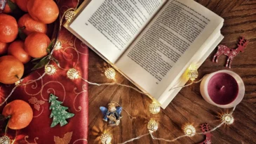 A book lays open on table. It's surrounded by oranges, Christmas lights and a candle.