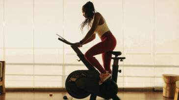 Young African woman engaging in indoor cycling on a stationary bike, using the smart equipment to follow a cardio routine. Focused and determined, she pedals in pursuit of her health and wellness goals.