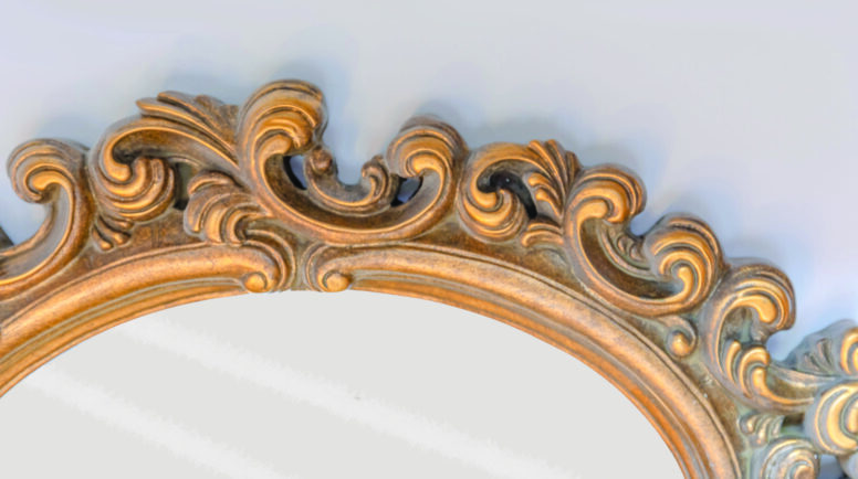 A partial view of an ellipse shaped old mirror with gold coloured and ornamented frame on a white wall. Posted with "Sometimes I wish I had a face of my own."