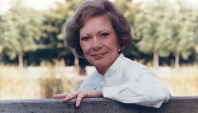 A 1993 photo of Rosalynn Carter sitting on a bench, trees in the background.