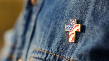 Close up of Christian cross pin with American flag colors is pinned on blue jeans jacket. Patriotism and religious rights concept