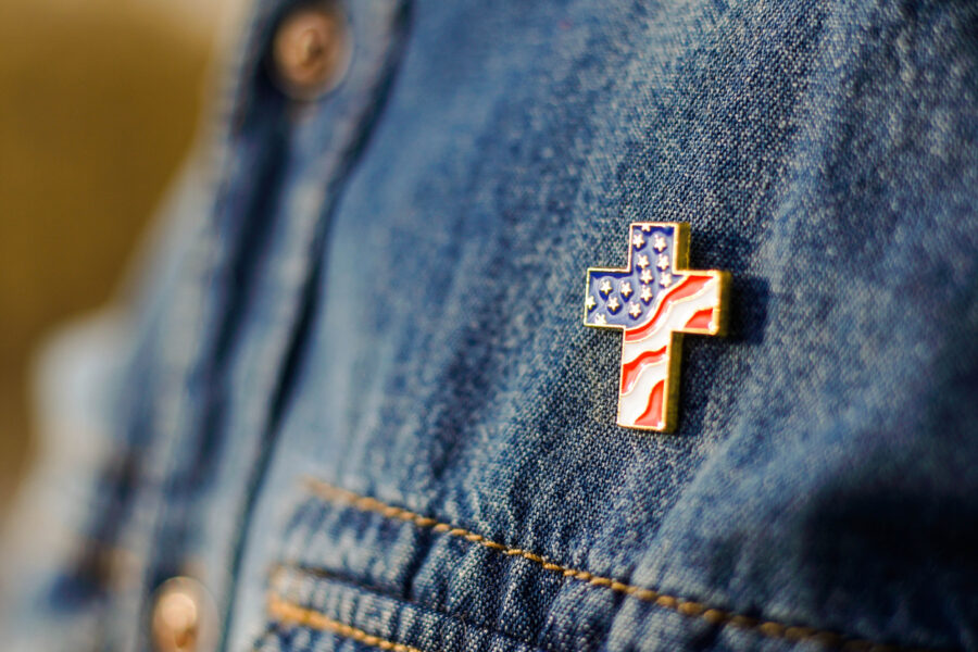 Close up of Christian cross pin with American flag colors is pinned on blue jeans jacket. Patriotism and religious rights concept