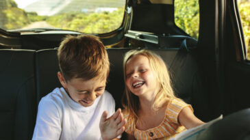 Small boy and girl sitting on backseat of car looking at map and smiling. Kids traveling in a car on roadtrip playing with a map.