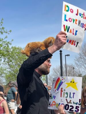A man stands at a protest with a stuffed animal on his head.