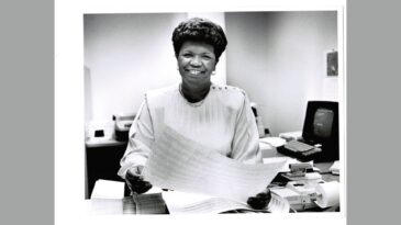 A black and white photo of a woman smiling while working at a desk.