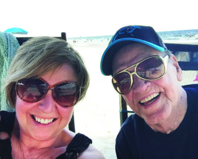 A selfie of a woman and her father sat beside each other smiling on a beach.