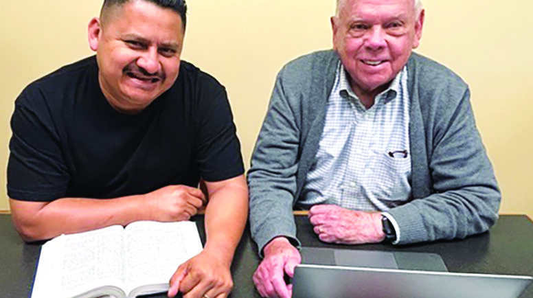 Two men sat at a table smiling with a book and laptop.