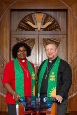 Two pastors wearing green stoles pose by a baptismal font