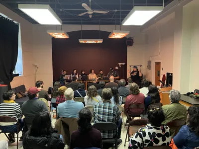 A panel sits on stage while a room full of people sitting on folding chairs listens.