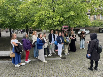 A group of college students stand on a stone-lined street in Scotland. There are cars in the background and trees lining a street.