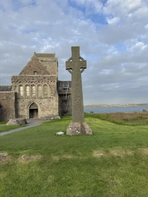 An old, stone Celtic Cross statue sits in front of an old, stone church. The grass around the statue and church is bright green and there is water in the background. 