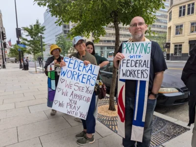 A group of four people stand outside with signs. Some of them wear stoles. One sign says, "Praying with federal workers." The other sign says, "Federal workers: we see you; we appreciate you; we pray with you."