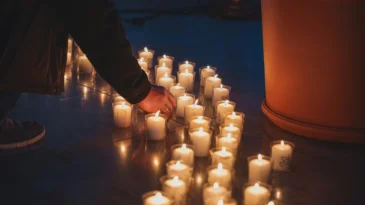 several rows of votive candles line the ground. A hand reaches down to light one.