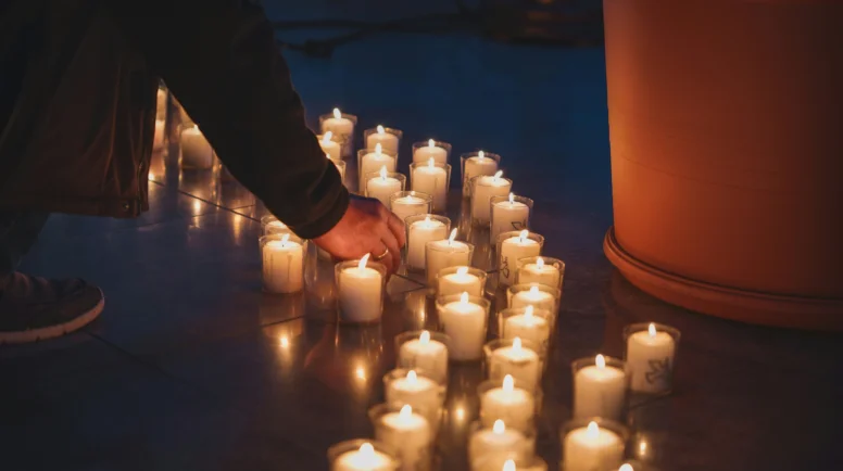 several rows of votive candles line the ground. A hand reaches down to light one.