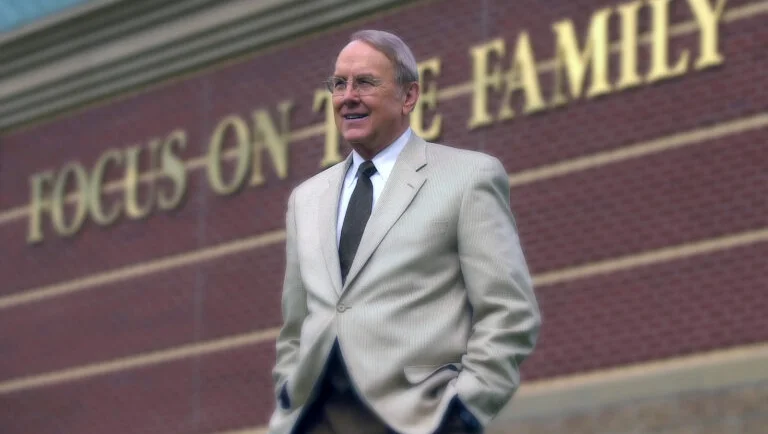 A white man with gray hair stands in a light blazer and tie outside of a building with the words "Focus on family" on the side.