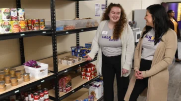 Two Converse students stand by food pantry shelves and talk to each other.