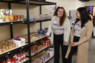 Two Converse students stand by food pantry shelves and talk to each other.