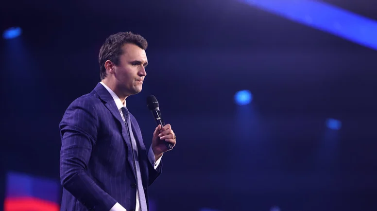 Charlie Kirk stands on stand in a blue suit holding a microphone, looking pensive.