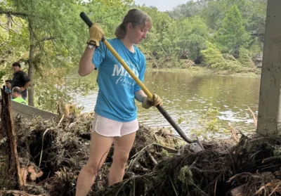 A young adult stands on top of a pile of water logged organic matter. There's a river in the background and the staff member is holding a shovel.