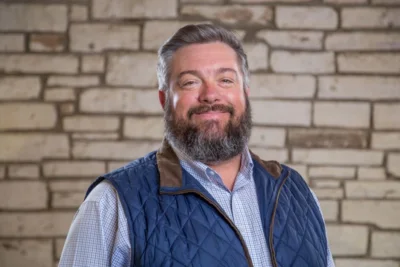 A man with a beard smiles at the camera. He's wearing a blue button down and a blue vest.