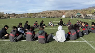 Students sit in a large circle on a football field. They're stretching and wearing athletic uniforms.