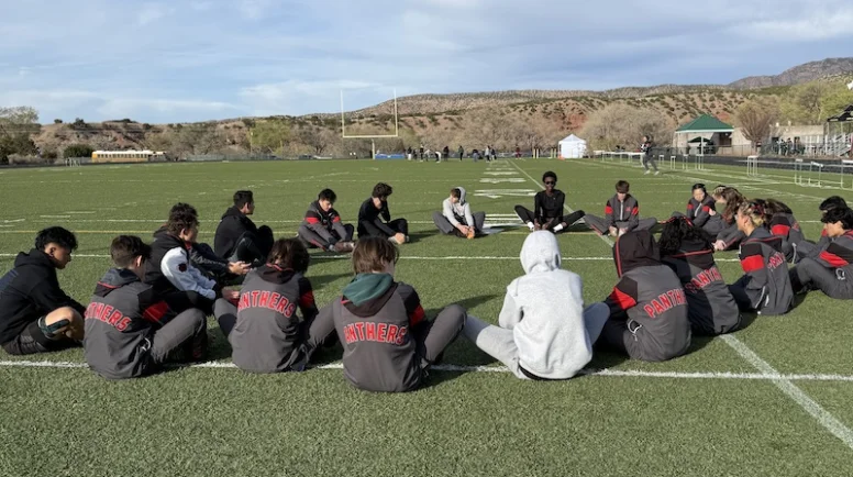 Students sit in a large circle on a football field. They're stretching and wearing athletic uniforms.