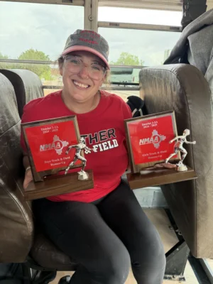 Coach Essie sits on a school bus wearing school colors and holding two trophies.