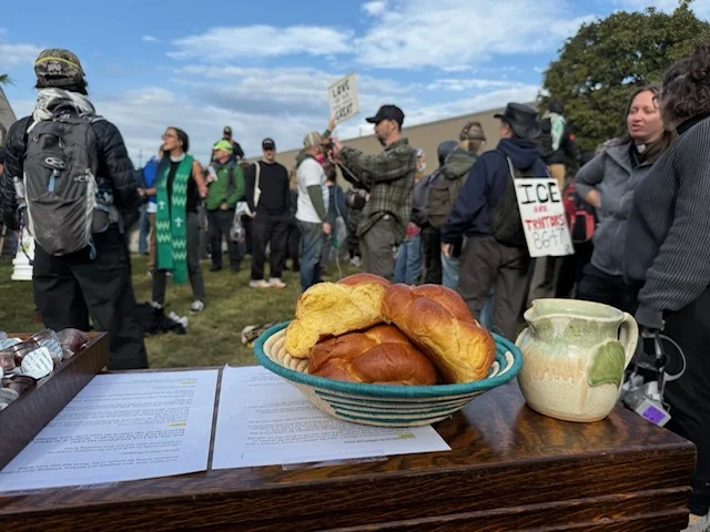 Challah bread, individual communion cups, and a clay jug containing grape juice sit on a table outside. In the background, protestors hold signs and speak to one another.