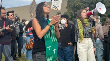A woman in a clerical collar and green stole, Rev. Emily Heitzman, stands in the foreground of in a small crowd of people.