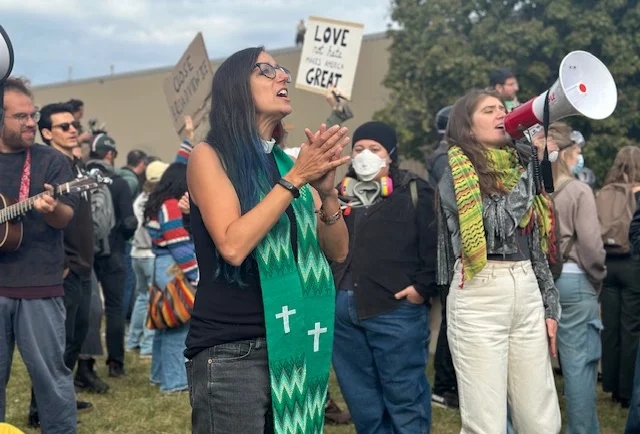 A woman in a clerical collar and green stole, Rev. Emily Heitzman, stands in the foreground of in a small crowd of people.