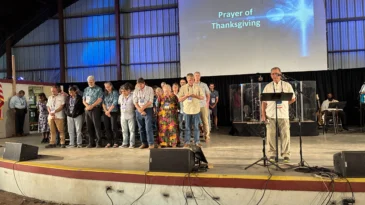 A man prays a prayer of Thanksgiving over a group of peole standing on a stage.