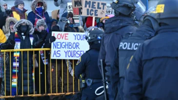 Police stand in front of protesters, including one holding a sign "love your neighbor as your self"