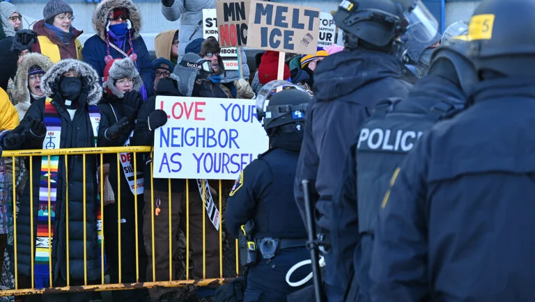 Police stand in front of protesters, including one holding a sign "love your neighbor as your self"
