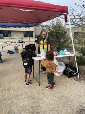 A woman in a stole stands under a canopy tent speaking to two children, whose backs are facing the camera.