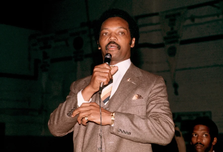 Jesse Jackson Sr. stands wearing a brown suit and holding a microphone.