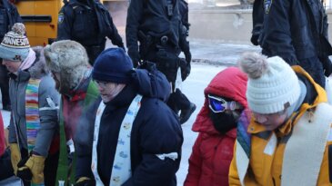 A group of protesters, many wearing stoles, wear cold weather gear and kneel outside of the Minneapolis airport.