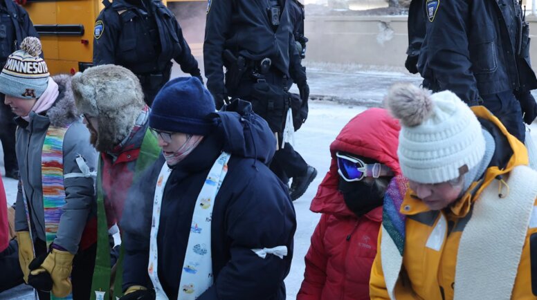 A group of protesters, many wearing stoles, wear cold weather gear and kneel outside of the Minneapolis airport.