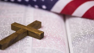 A holy Christian cross laying on an open Bible with an American flag in the background.