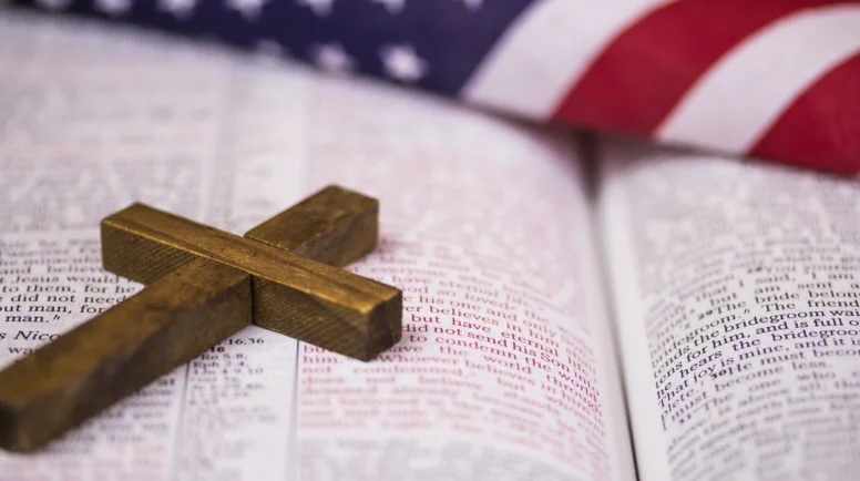 A holy Christian cross laying on an open Bible with an American flag in the background.