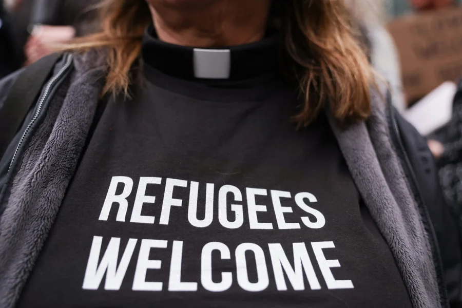 A woman wears a clerical collar and a tshirt that says "Refugees welcome"