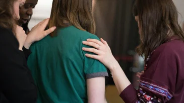 Three women standing. The woman in the middle is being prayed over by other two women.
