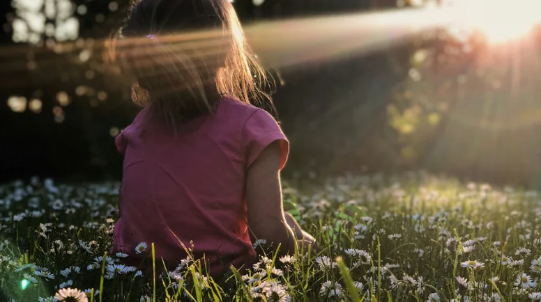 Photo of a girl sitting in a field of flowers. The sun is shining on her face.