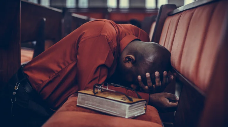 A man bent over in a posture of prayer, his bible and eye glasses next to him on the pew.