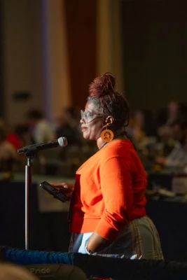 A woman in orange stands behind a microphone. 
