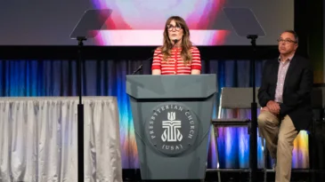 A woman stands behind a podium with the PC(USA) logo.