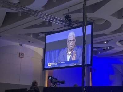 A photo of a woman speaking, displayed on a large screen in a convention hall. 