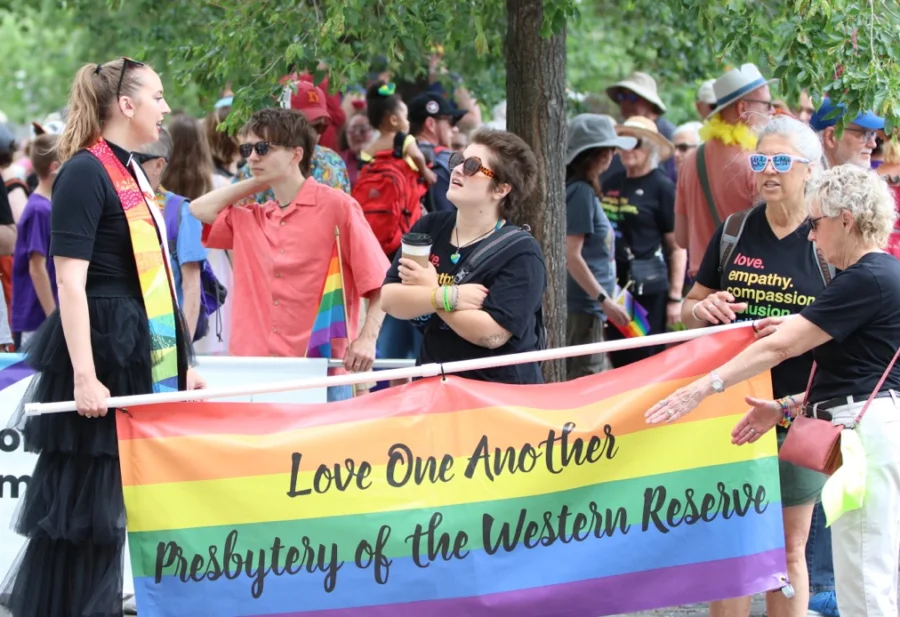 A woman in a clerical collar holds a rainbow flag with the words "love one another/ Presbytery of the Western Reserve"