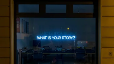 Window of a dark room with a light up sign saying "what is your story?".
